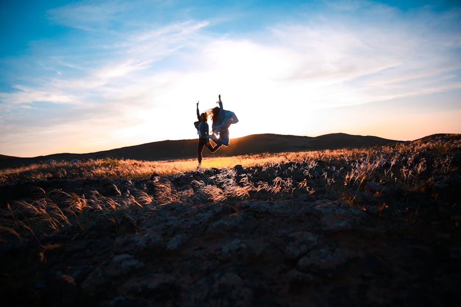 Two friends jump joyfully at sunset in a rocky field, creating a silhouette against the vibrant sky.